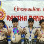 Four smiling schoolchildren in beige uniforms pose with colorful handmade badges in front of a banner that reads observation of